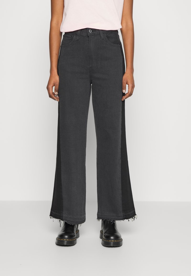 Personne portant un jean denim noir à jambes larges avec des ourlets effilochés, un haut rose clair rentré et des bottines noires.