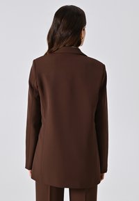 Blazer marrone realizzato in tessuto liscio, caratterizzato da una vestibilità sartoriale, rever a scialle e maniche lunghe, visto da dietro.