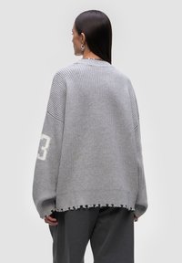 Szary, dziany sweter z prążkowaną teksturą, w oversize'owym fasonie, z surowym wykończeniem. Posiada biały nadruk „3” na lewym rękawie oraz okrągły dekolt.