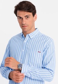 Camisa de botones a rayas en azul claro con un bolsillo en el pecho, que presenta bordado rojo. La tela parece suave con un cuello estructurado.