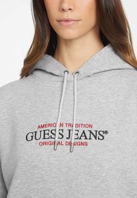 Hanorac gri cu text brodat în roșu și negru: "TRADIȚIE AMERICANO-ORIGINALĂ GUESS JEANS DESIGNURI. Detaliu cu șnur pentru glugă.