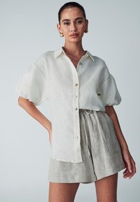 Witte linnen overhemd met korte mouwen en een knoopsluiting aan de voorkant, versierd met een stervormig accent, gecombineerd met beige linnen shorts met trekkoord.