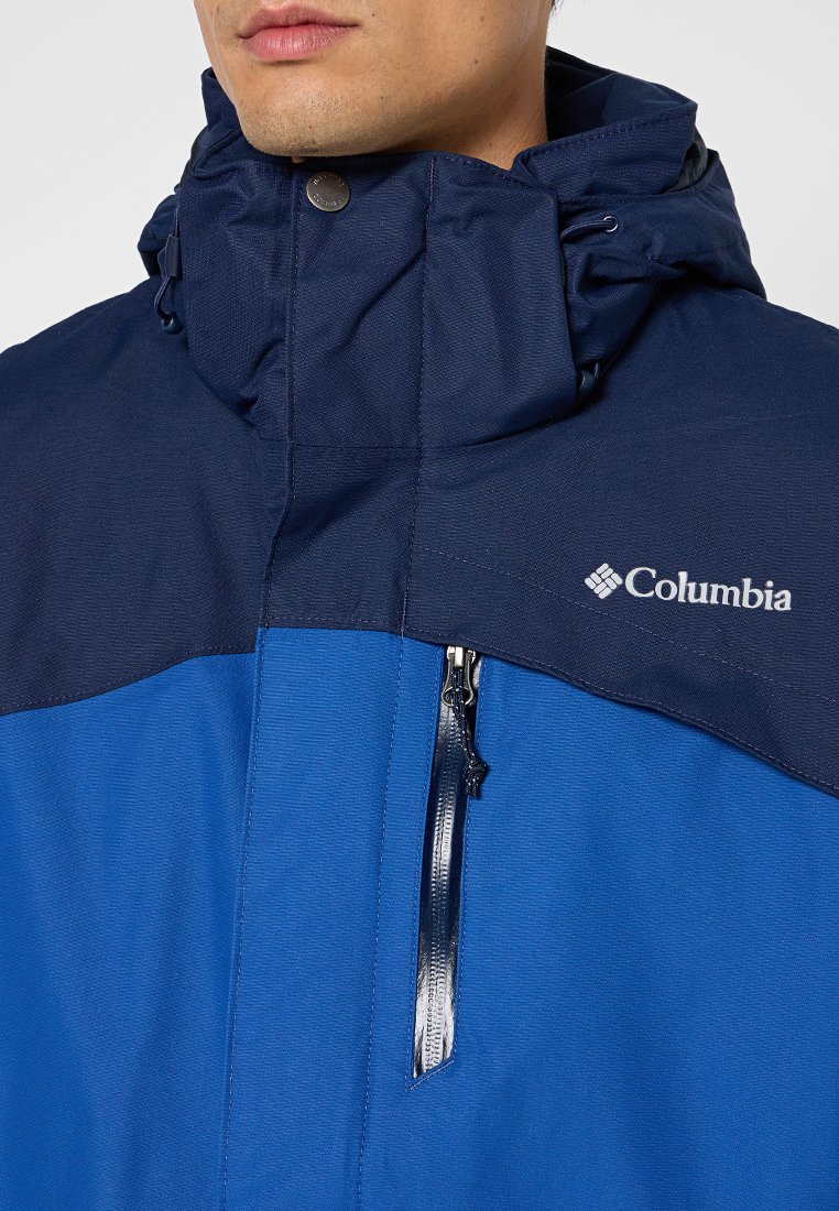 Blauwe waterdichte jas met een hoge kraag, een ritszak en het Columbia-logo op de borst. Het materiaal heeft een gestructureerde afwerking.