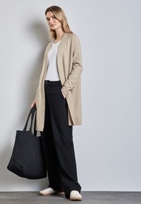 Beige lange cardigan van gestructureerd materiaal, gedragen over een wit shirt en zwarte wijde broek, met een zwarte gewatteerde tas en witte sneakers.