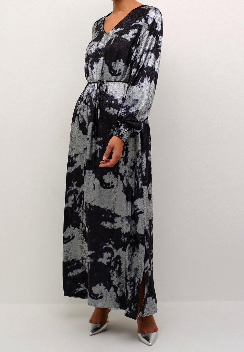 Femme portant une longue robe tie and dye noire et grise avec des manches longues, un col en V, une ceinture à la taille, une fente latérale et des talons pointus argentés.