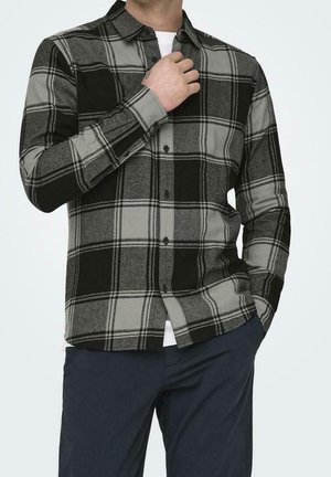 Homme portant une grande chemise à carreaux noire et grise boutonnée sur un sous-vêtement blanc avec un pantalon foncé, une main ajuste le col.
