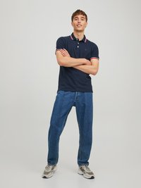 Námořnická polo košile s červeným a bílým pruhovaným límečkem, v kombinaci s volně střiženými modrými džíny a šedými teniskami, stojící proti neutrálnímu pozadí.