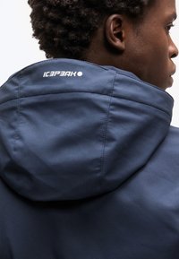 Κοντινή λήψη ενός ατόμου που φοράει μπλε μπουφάν με κουκούλα, με το λογότυπο «ICEPEAK» ορατό στο πίσω μέρος της κουκούλας.