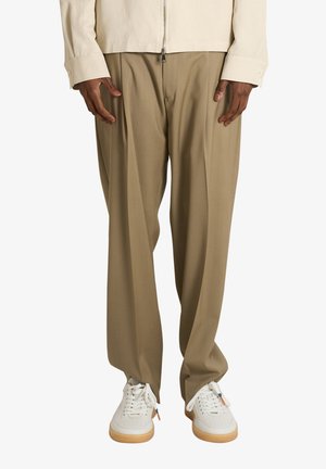 Persona che indossa pantaloni beige larghi con pieghe frontali abbinati a scarpe da ginnastica bianche con suola in gomma, in piedi su sfondo bianco.