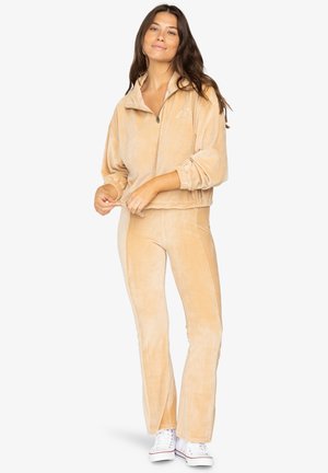 Tan velour træningsdragt med lynlåsjakke og tætsiddende bukser. Tekstureret stof, ribbet design og manchetter på ærmerne. Bæres med hvide sneakers.