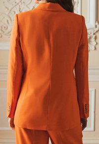Blazer sur mesure orange avec revers crantés, silhouette élégante et poignets boutonnés. Le tissu semble lisse avec un design structuré.