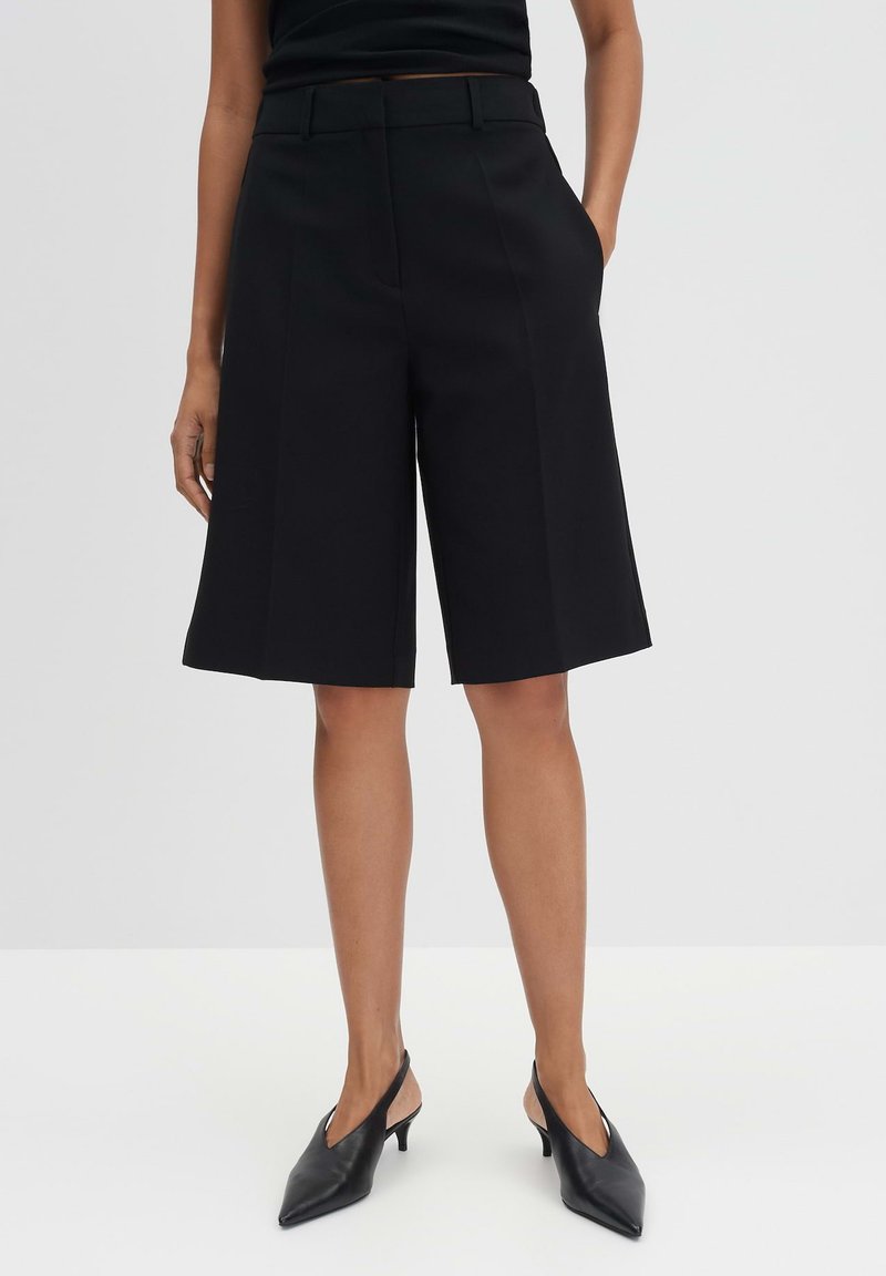 Schwarze maßgeschneiderte Shorts mit hoher Taille, gefalteter Front und Seitentaschen. Getragen mit schwarzen Slingback-Absätzen, die ein spitz zulaufendes Design aufweisen.