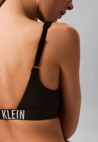 Prim-plan al spatelui superior al unei femei care poartă un sutien negru cu nervuri, cu bretele reglabile și inscripția „KLEIN” pe bandă, pe un fundal gri.