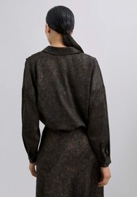 Blusa marrón con una tela texturizada, de mangas largas y cuello. La parte trasera presenta un corte holgado y un patrón sutil, con puños abotonados.