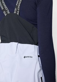 Sive in svetlo modri GORE-TEX smučarski kombinezoni z nastavljivimi naramnicami, oblečeni čez temno modro dolgi rokav, z več žepi.
