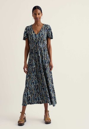 Vrouw staand in een marineblauwe midi-jurk met bloemenprint, korte mouwen, V-hals en bruine platformzandalen tegen een effen achtergrond.