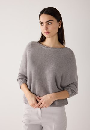 Young woman with long dark hair wearing a loose gray knit sweater and light gray pants, looking sideways with hands clasped in front.