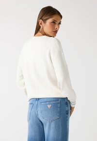 Maglione bianco con una vestibilità morbida e polsini a coste, abbinato a jeans in denim azzurro chiaro con un piccolo badge logo sulla tasca posteriore.