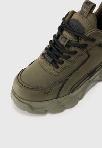 Sneaker verde oliva con materiale sintetico testurizzato, linguetta imbottita e lacci neri. Suola in gomma spessa con dettagli arrotondati.