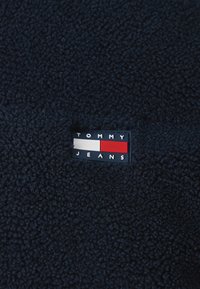Marinefarget fleece-stoff med en teksturert overflate. En merkelapp med "TOMMY JEANS" i hvite og røde blokker er tydelig vist.