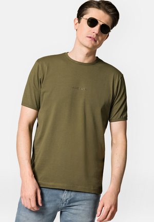 Oliwkowy t-shirt z krótkim rękawem, okrągłym dekoltem i subtelnym detalem logo na klatce piersiowej. Połączony z jasnoniebieskimi dżinsami.