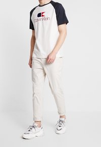 Camiseta blanca de algodón con mangas raglán en navy y logo de Calvin Klein, combinada con pantalones beige ajustados y zapatillas blancas con acentos grises.