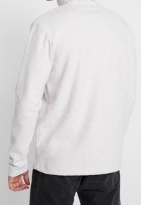 Suéter de punto gris claro con rayas verticales texturizadas, cuello alto, mangas largas y aberturas laterales para mayor libertad de movimiento.