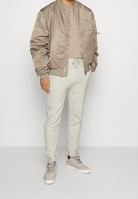 Ljust brunt bomberjacka med satinfinish, beige t-shirt, ljusgrå sweatpants och bleka grå sneakers med mockadetaljer.