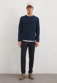 Marineblauw textuurtrui, wit overhemd met zwarte strepen eronder, zwarte broek, tan schoenen en een lichtbeige pet.