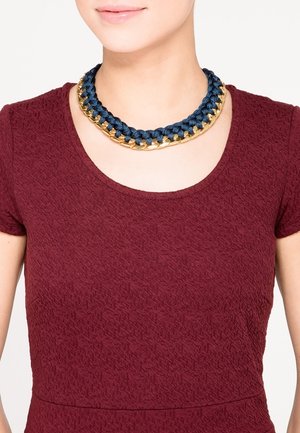Femme portant une robe rouge foncé texturée et un collier épais avec du tissu bleu marine entrelacé et des maillons de chaîne dorés.