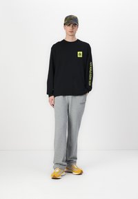 Long-sleeve black t-shirt with neon yellow logo and text, paired with gray sweatpants, yellow and gray sneakers, and a camo cap.