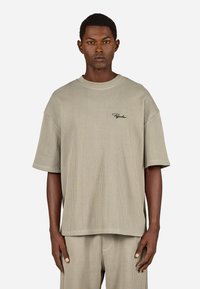 Mężczyzna w beżowej, prążkowanej koszuli z krótkim rękawem i pasujących spodniach, z czarnym haftem na piersi i kieszeni spodni, stoi na tle białym.