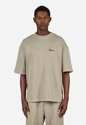 Mann in beigem, geripptem Kurzarmhemd und passender Hose mit schwarzem Sticktext auf der Brust und der Hosentasche, vor einem weißen Hintergrund stehend.