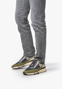 Henkilö, joka on pukeutunut harmaisiin farkkuihin ja vihreisiin, mustiin ja beigeihin lenkkareihin, seisoo valkoista taustaa vasten.