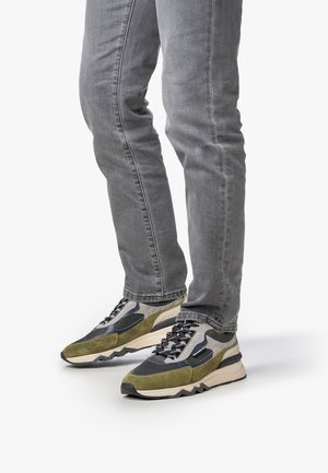 Persona che indossa jeans grigi e scarpe da ginnastica verdi, nere e beige in piedi su uno sfondo bianco.