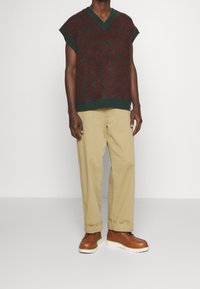 Grön och röd mönstrad stickad väst med V-ringning, tillsammans med ljusbeige byxor och bruna skor med vit sula.