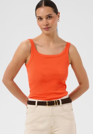 Kvinde med mørkt hår i en hestehale iført en orange ærmeløs top, cremefarvede bukser med et mørkebrunt bælte, stående med hænderne bag ryggen.