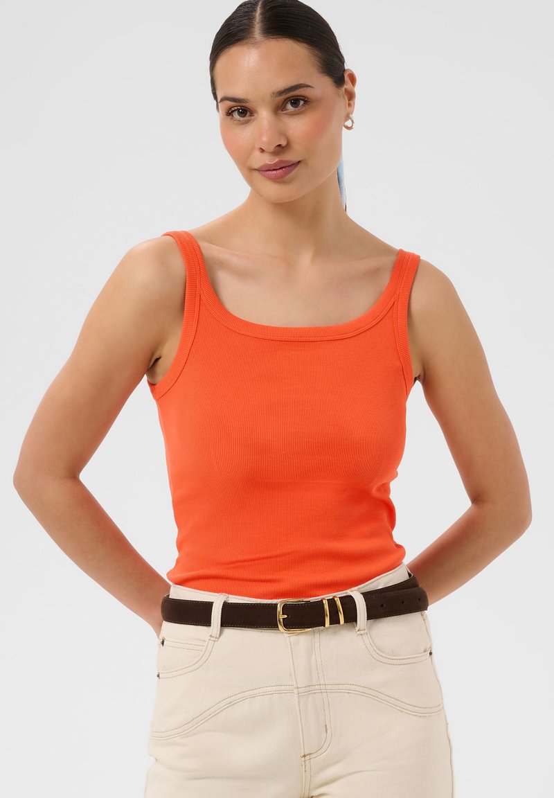 Femme aux cheveux foncés en queue de cheval, portant un haut orange sans manches, pantalon crème avec une ceinture marron foncé, debout, les mains dans le dos.