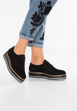Schwarze Schnür-Plateauschuhe kombiniert mit hochgekrempelten blauen Jeans mit schwarzer Blumenstickerei an den unteren Beinen.
