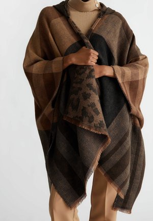 Personne portant un poncho à motif brun sur un col roulé beige et un pantalon beige, tenant les bords du poncho fermés au niveau de la poitrine.