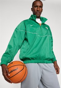Mann hält Basketball, trägt eine grüne Zip-Up-Jacke mit Sportlogo und hellgraue Shorts, steht vor einem schlichten Hintergrund.