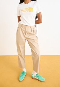 Camiseta blanca con logo amarillo, pantalones beige con pliegues y zapatillas verde azulado con detalles blancos sobre un fondo amarillo.