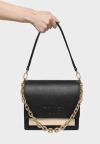 Mano sosteniendo un bolso de cuero negro de TOUS con cadena dorada y acento beige sobre un fondo gris claro liso.