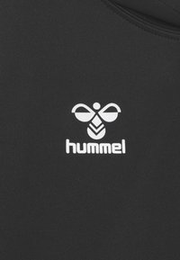 Logo d'abeille stylisé blanc avec des ailes et un corps géométriques au-dessus du nom de la marque "hummel" sur un fond en tissu noir.
