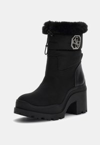 Bottine noire avec un talon épais, bord en fausse fourrure, cordon de serrage en haut et patte décorative en strass sur le côté extérieur.