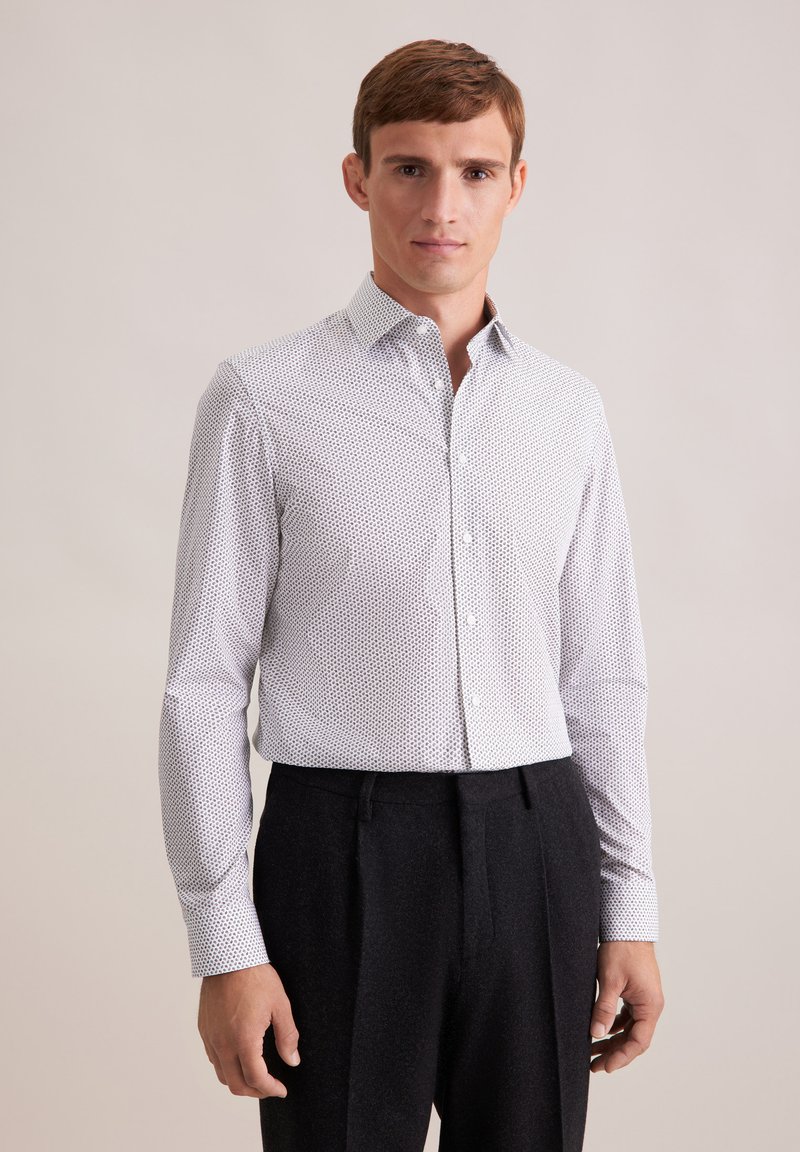 Camicia bianca con un delicato motivo a pois neri, colletto classico, maniche lunghe e vestibilità sagomata, abbinata a pantaloni scuri.