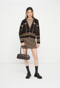 Braune Zip-Jacke mit kariertem Muster, kombiniert mit einem taillierten Rock in Braun und Schwarz. Eine dunkle Handtasche und knöchellange Socken runden den Look ab.