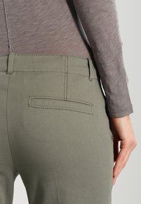 Grüne Hose mit glatter Textur, ausgestattet mit einer Gesäßtasche und einem Gürtelbund. Das Oberteil ist ein eng anliegendes, langärmliges, hellgraues Hemd.