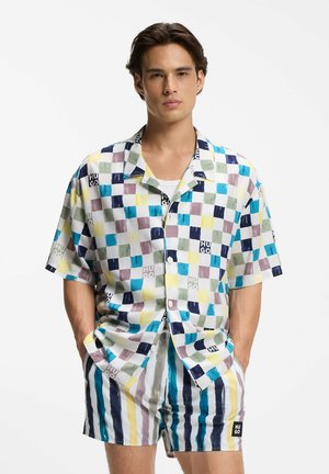 Młody mężczyzna ubrany w kolorową koszulę w kratę z krótkim rękawem oraz pasujące, paskowane szorty, stojący z rękami w kieszeniach na białym tle.