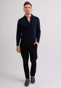 Navy modrá zipová pletená svetr, černé kalhoty a modré sportovní boty. Svetr má vysoký límec a hladkou texturu.